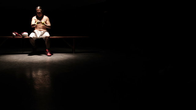 An older boxer sits on a bench in the shadows and wraps his hands