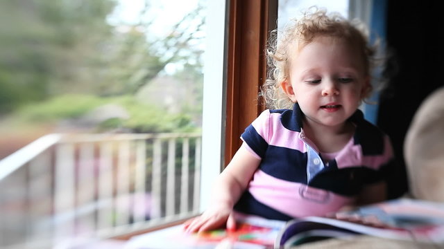 An adorable little girl sits by a window and flips through her book