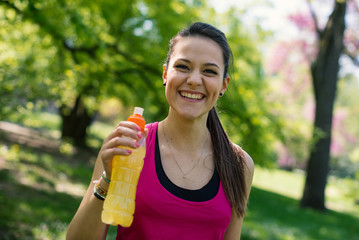 Smiling young woman holding energy drink outdoors in a park. - 84080121
