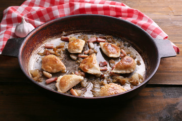 Fried dumplings with onion and bacon in frying pan, on wooden table background