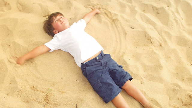 A family sits in the sand at the beach and watch a young boy makes sand angels