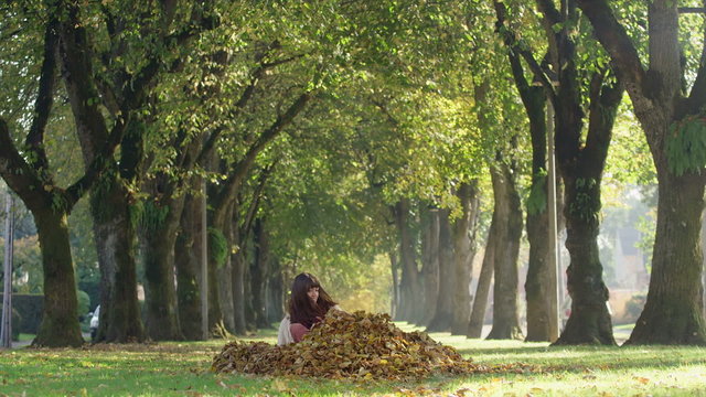 A young woman falls and lays down in a pile of dead leaves in the park, in slow motion