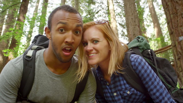 A man and a woman standing in the forest with backpacks, look into the camera and make funny faces