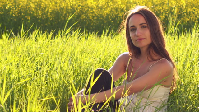 A cute young woman sits in the grass in an open field and looks at the camera