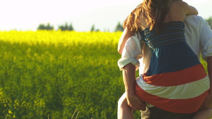 A young man gives a woman a piggyback ride a open field with yellow flowers