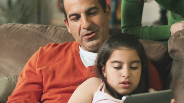 Mother looks over the couch on the father as he reads a tablet with his daughter