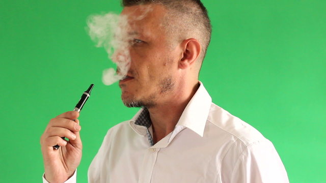 profile of man vapouring electric cigarette in front of green background. vapour (smoke) is visible. 