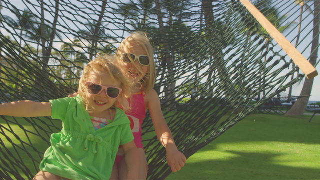Two young girls lay in a hammock, wearing sunglasses and smile into the camera