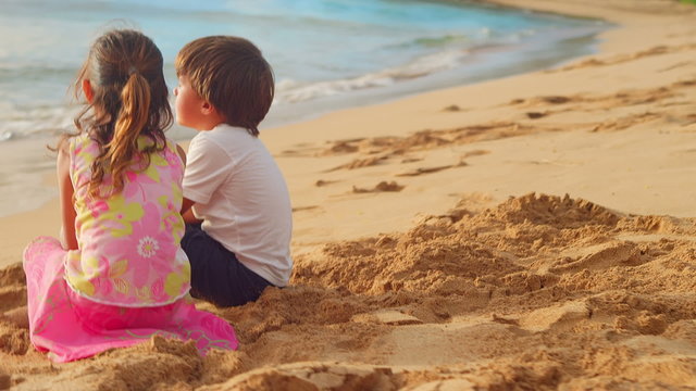 A young brother and sister with their arm around each other sit on a beach 