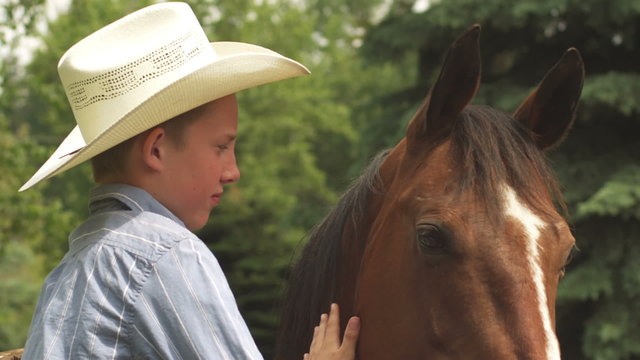 A boy in a cowboy hat sits on a fence with his horse and pets him