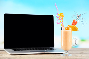 Laptop and glass of summer cocktail on wooden table, on bright blurred background