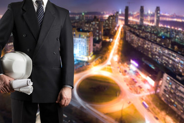 Architect hand holding white safety helmet for workers security and blueprints standing in front of blurred urban background