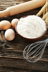 Bowl of wheat flour with eggs and whisk on brown wooden backgrou
