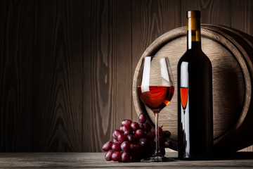 Black bottle and glass of red wine with grapes and barrel
