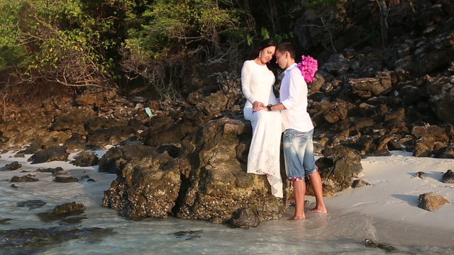 bride sits on rock and groom holds hands at sunrise	