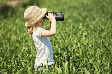Little blonde girl in straw hat looking through binoculars outdo