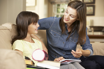 Mother Helping Daughter With Homework Sitting On Sofa At Home