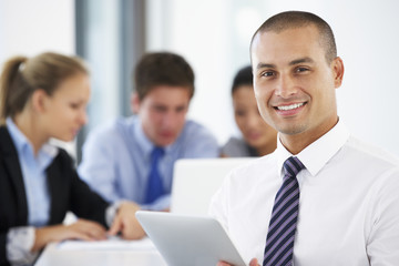 Portrait Of Male Executive Using Tablet Computer With Office Meeting In Background