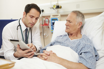 Obraz premium Doctor Sitting By Male Patient's Bed Using Digital Tablet