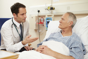 Doctor Sitting By Male Patient's Bed In Hospital