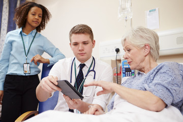 Obraz premium Doctor Sitting By Female Patient's Bed Using Digital Tablet