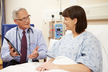Fototapeta premium Doctor Sitting By Female Patient's Bed Using Digital Tablet