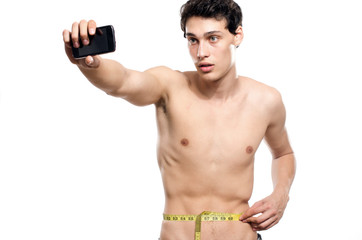 Skinny man taking a selfie with his phone while measuring his waist with a centimeter. Beautiful teenager taking a photo for facebook.Anorexic young man training to become stronger