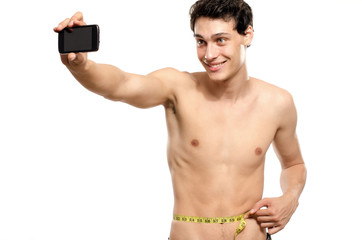 Fototapeta premium Skinny man taking a selfie with his phone while measuring his waist with a centimeter. Beautiful teenager taking a photo for facebook.Anorexic young man training to become stronger