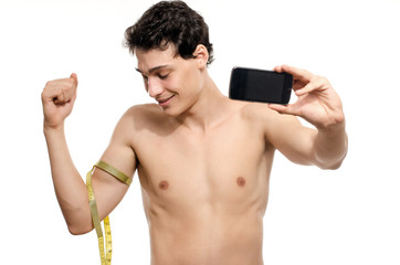 Skinny man taking a selfie with his phone while measuring his biceps with a centimeter. Beautiful teenager taking a photo for facebook.Anorexic young man training to become stronger