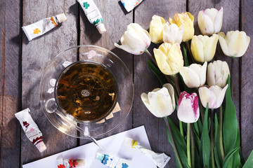 Cup of herbal tea with tulips and colorful paint in tubes on wooden table, top view