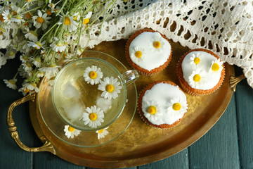 Cup of chamomile tea with chamomile flowers and tasty muffins on tray, on color wooden background
