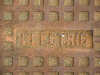 A rusty manhole cover with the word Electric on it, describing what utilities the cover gives access to.