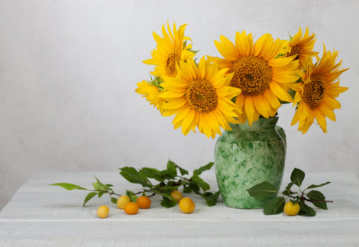 Bouquet of sunflowers in old ceramic jug against a white wooden wall. In the foreground branches with ripe cherry plum