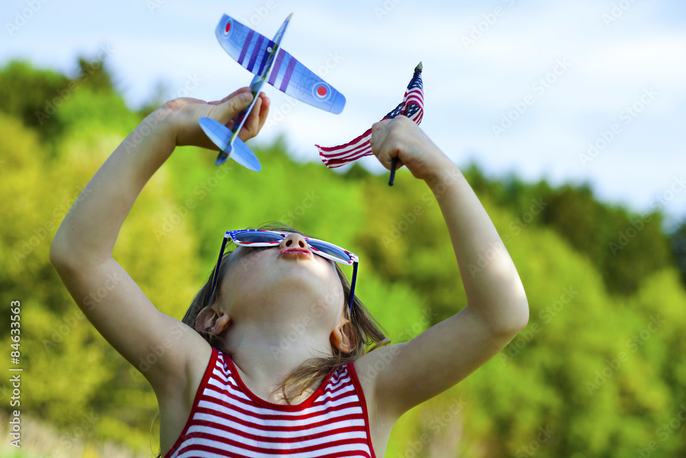 Wall mural patriotic girl playing with airplane while holding american flag outside