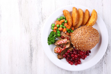 Beef with cranberry sauce, roasted potato slices, vegetables and bun on plate, on color wooden background