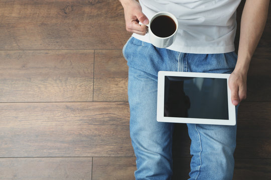 Young Man Sitting On Floor With Tablet And Cup Of Coffee In Room