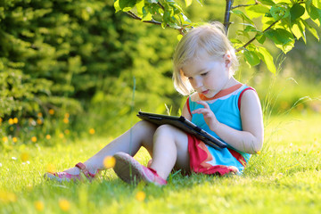 Happy preschooler child playing with tablet pc sitting in the garden on the grass. Cute little girl relaxing outdoors in the park enjoying sunny summer day. Young generation leisure.