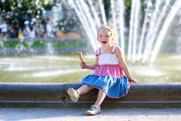 Happy child enjoying summer day outdoors. Cute little toddler girl eating ice cream outdoors in city park. Beautiful blurred fountain at background.