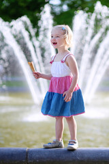 Naklejka premium Happy child enjoying summer day outdoors. Cute little toddler girl eating ice cream outdoors in city park. Beautiful blurred fountain at background.