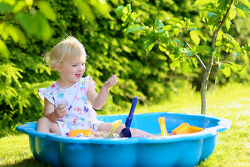 Happy little child, adorable blonde toddler girl having fun playing outdoors in the garden with plastic toys sitting in blue sand box on a sunny summer day