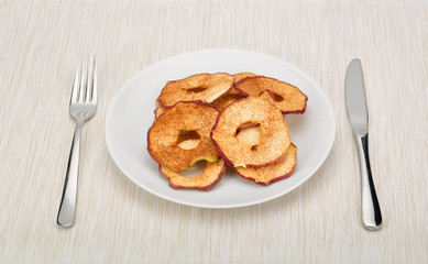 Dried apple slices sprinkled with cinnamon served on a white plate on tablecloth. Cutlery on both sides. 
