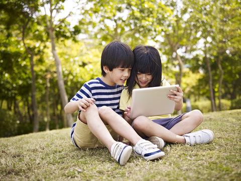 two asian children sitting on grass using tablet computer