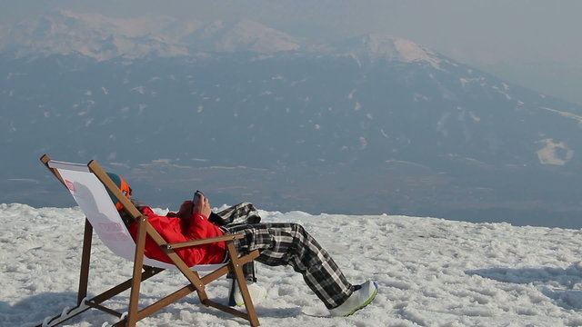 Snowboarder having rest in chair with gadget on top of mountain