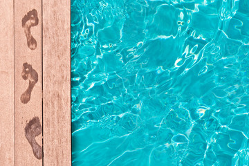 Top view poolside photo, wet footprints on the wooden deck of a swimming pool, blue clear water, summer holiday background with copy space