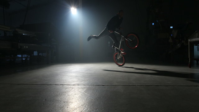 BMX rider doing tricks in dark warehouse.