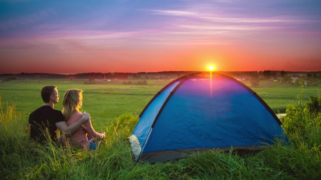The couple sit near camping tent by sunset (sunrise) background