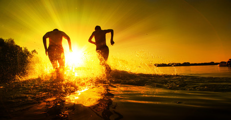Glückliche junge Menschen laufen und springen am See beim Sonnenuntergang