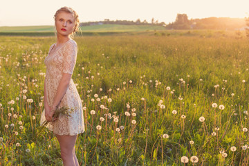 beautiful tender sweet girl in a white lace dress with a scythe on his head standing barefoot in a field of dandelions in the sunset