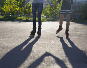 Fototapeta premium Female roller skaters holding hands. Body two female silhouette shadow on paved road skating roller skates holding hands of each other back-light sun urban background