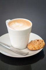 Cappuchino or latte coffe in a white cup and a biscuit on a dark grey background
